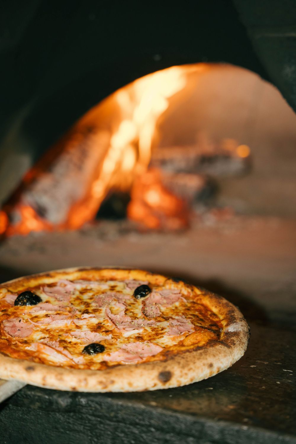 Pizza fraîchement cuite avec du jambon et des olives noires sur une peau devant un four à bois avec des flammes incandescentes.