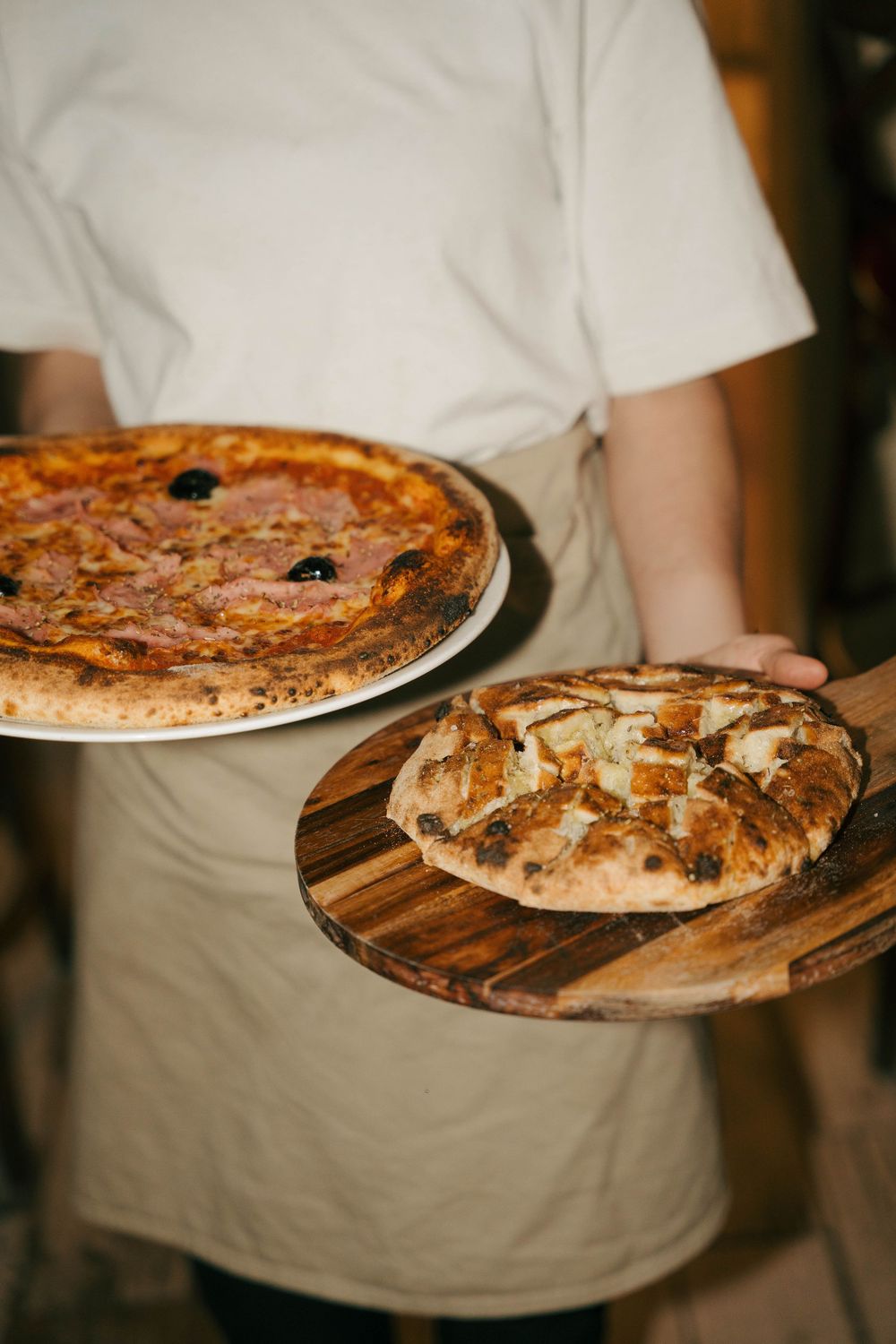 Chef tenant deux pizzas napolitaines fraîchement cuites sur des planches de bois, l'une garnie de salami et d'olives.