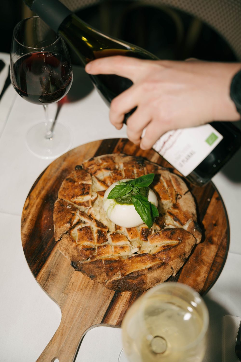 Pain rustique avec burrata et basilic sur planche de bois, servi avec du vin rouge versé à la table du restaurant.