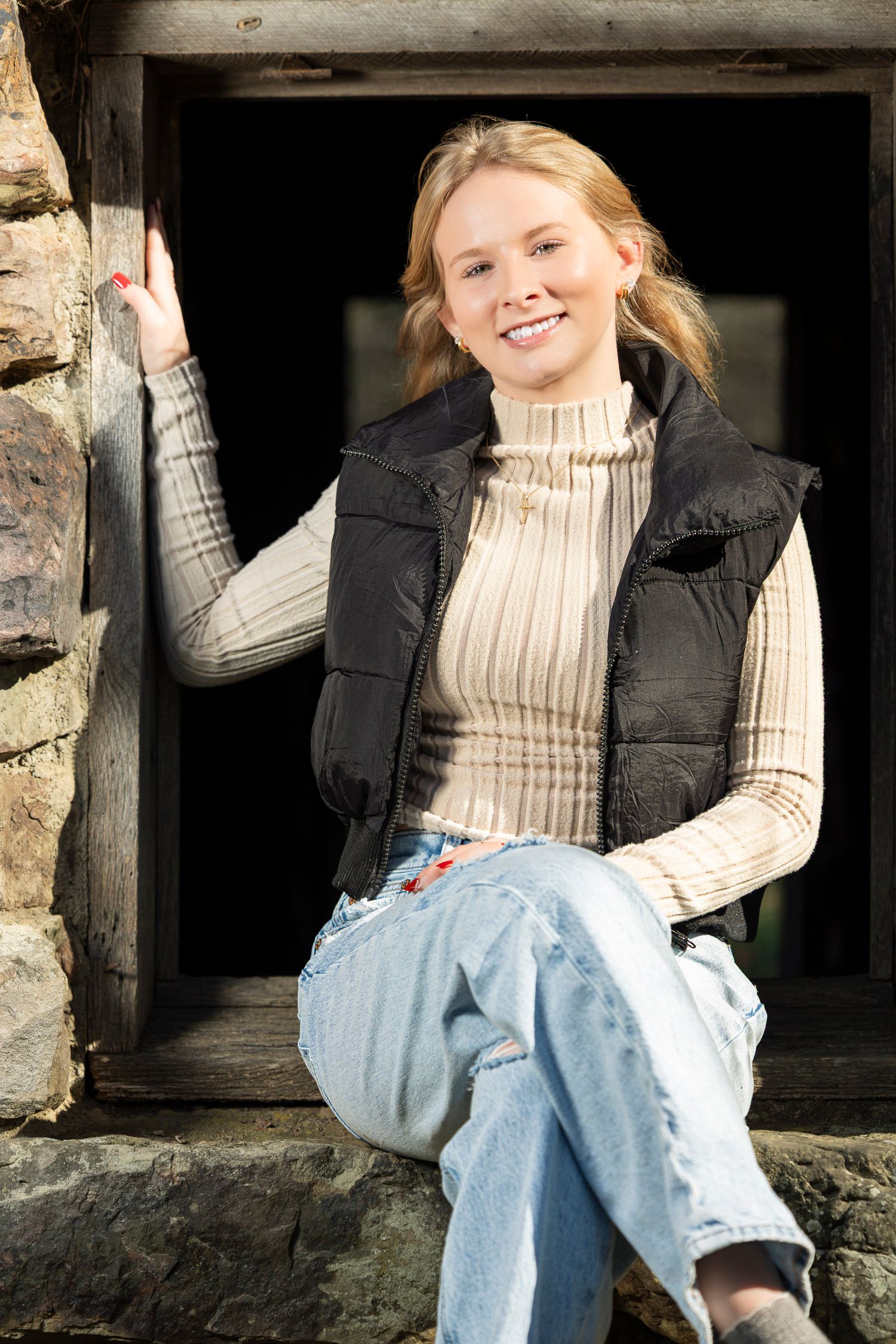 Smiling blonde woman in black vest and jeans sitting in rustic stone window frame outdoors.