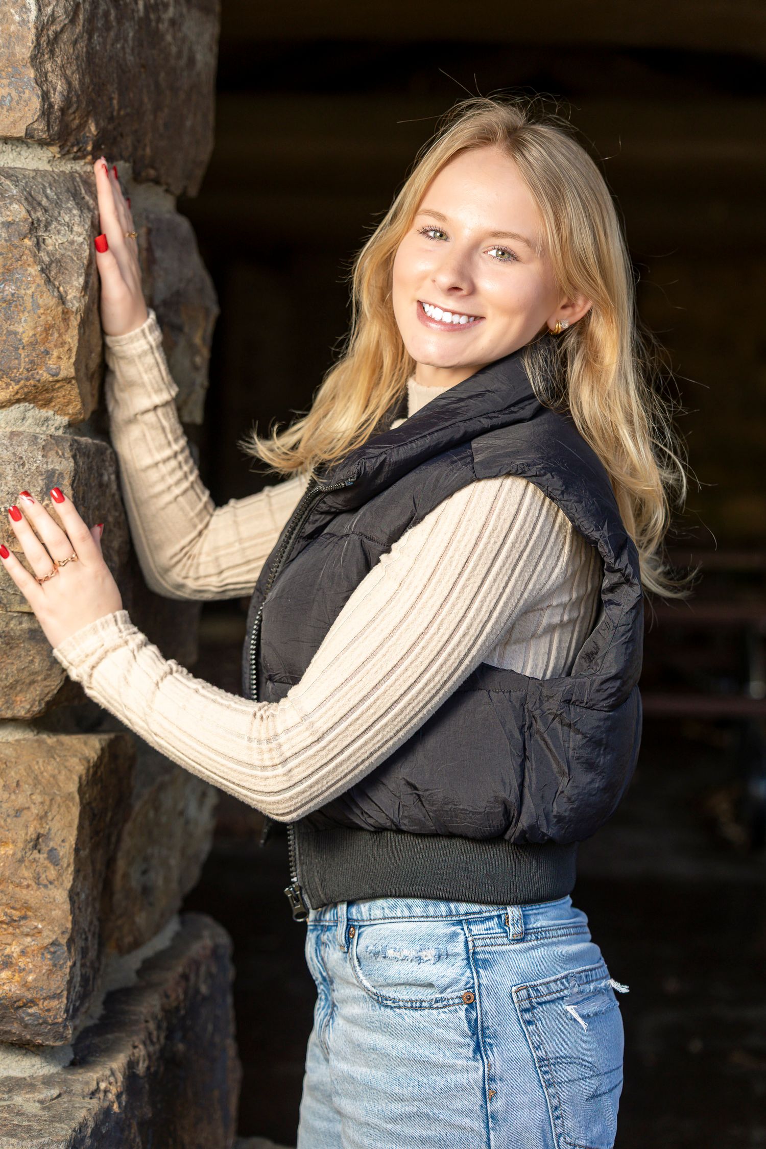 Smiling blonde woman in black vest and jeans leaning against a stone wall outdoors in natural light.