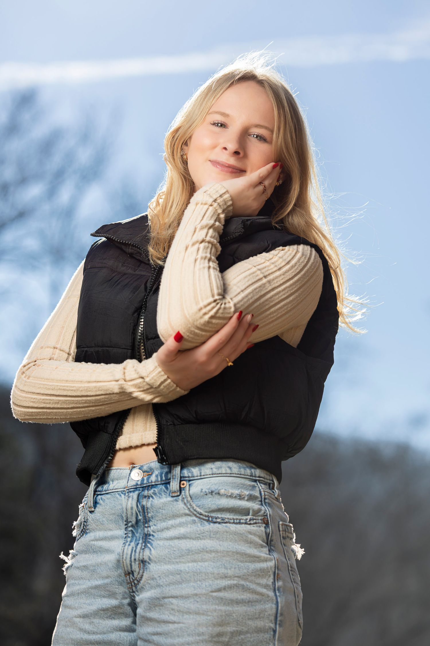 Young blonde woman in black vest and cream sweater poses outdoors against blue sky, smiling warmly.