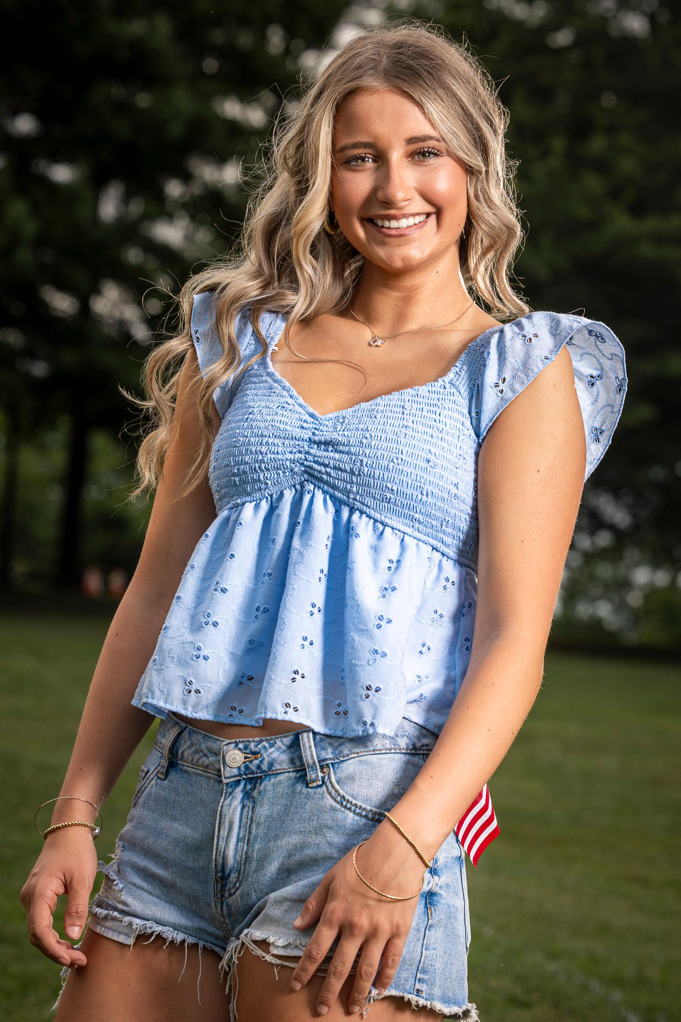 Young blonde woman smiling in blue eyelet top and denim shorts outdoors in a park setting.