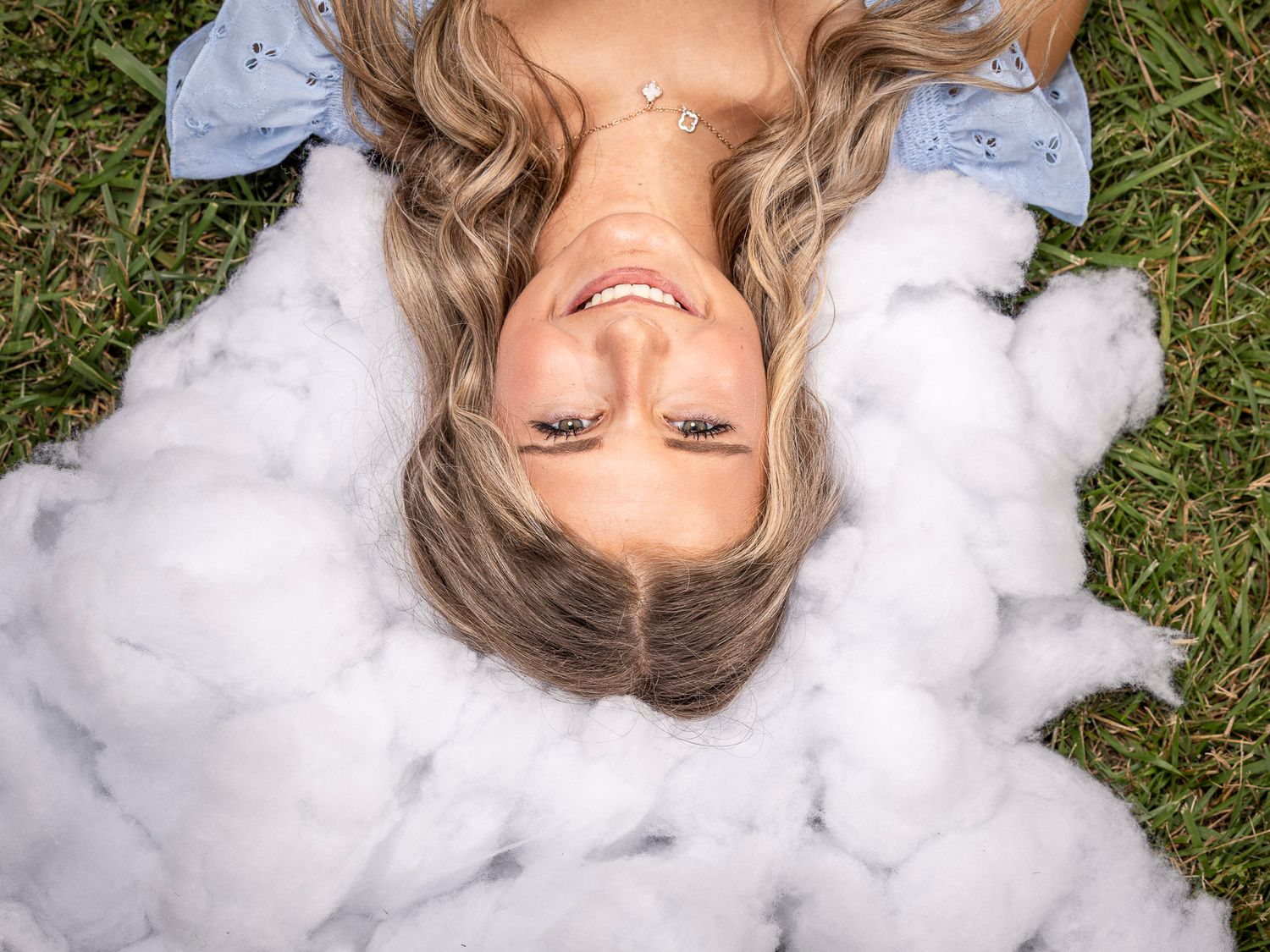 Smiling blonde woman lying on green grass surrounded by fluffy white clouds, viewed from above.