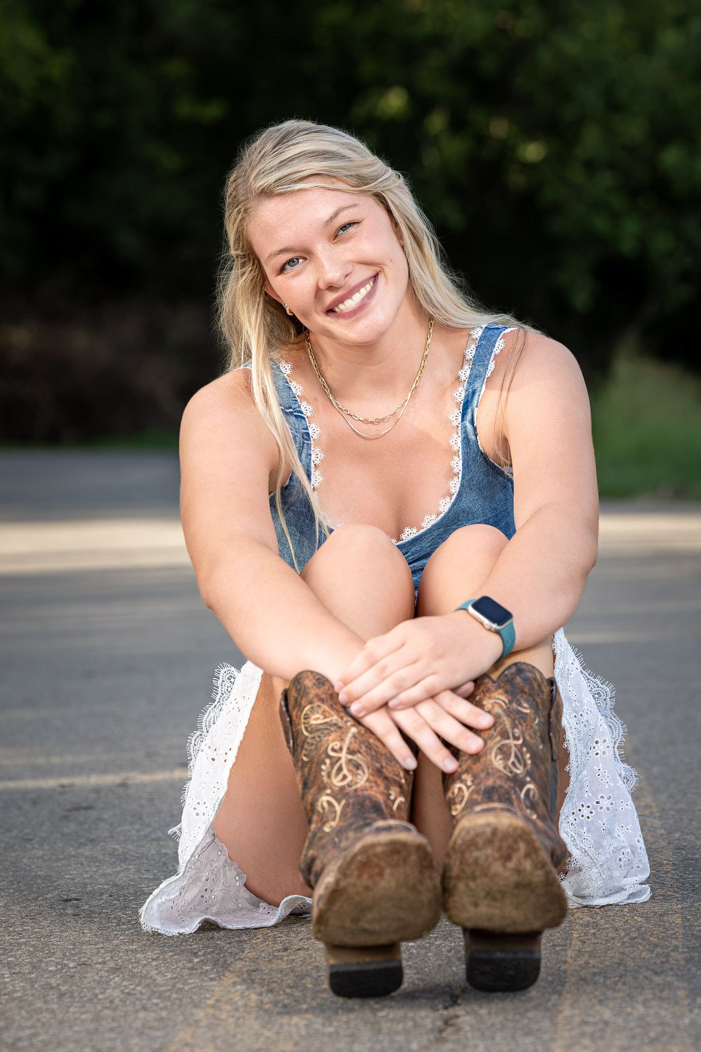 Smiling blonde woman sitting on pavement wearing denim top, white lace skirt, and brown cowboy boots outdoors.
