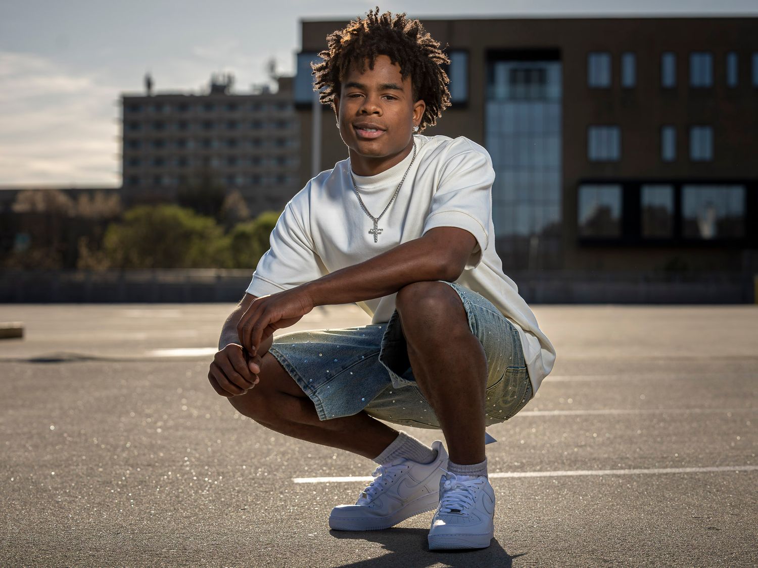 Young man in white tee and denim shorts crouching in urban parking lot, wearing white sneakers and gold chain.