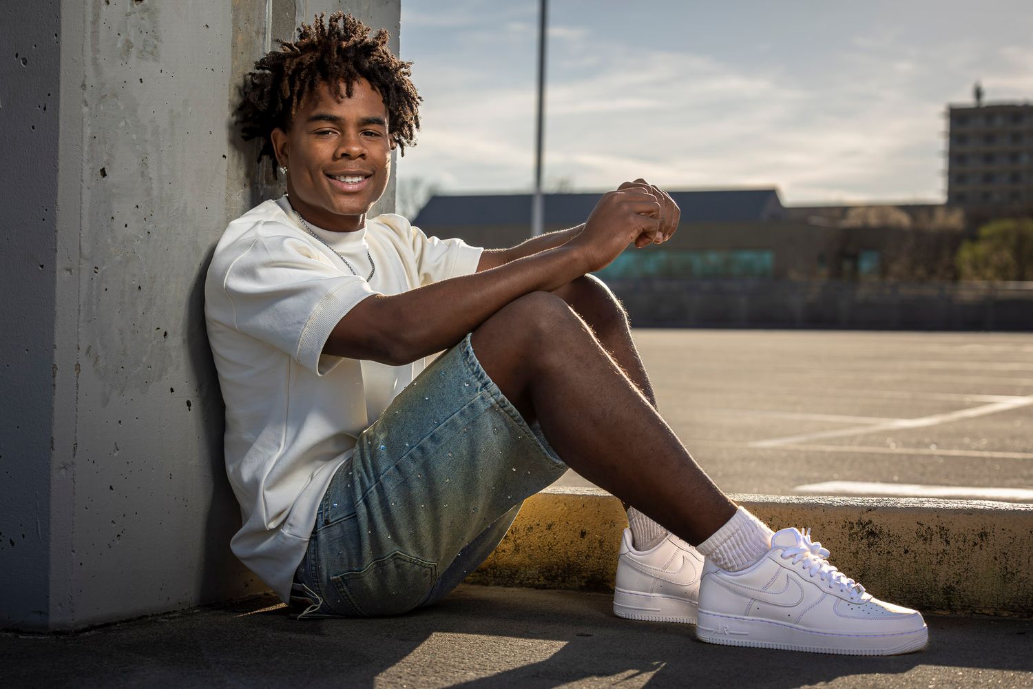 Young man in white tee, denim shorts, and Nike Air Force 1s sitting against a concrete wall outdoors.