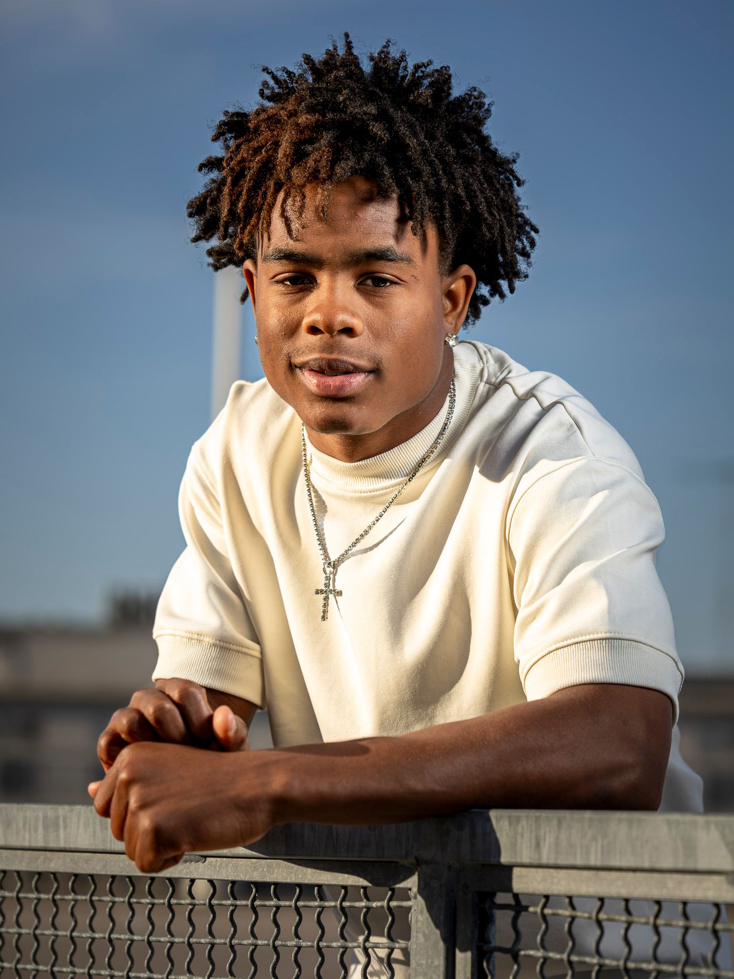 Young man with locs wearing cream hoodie leans on fence, silver cross necklace, blue sky background.