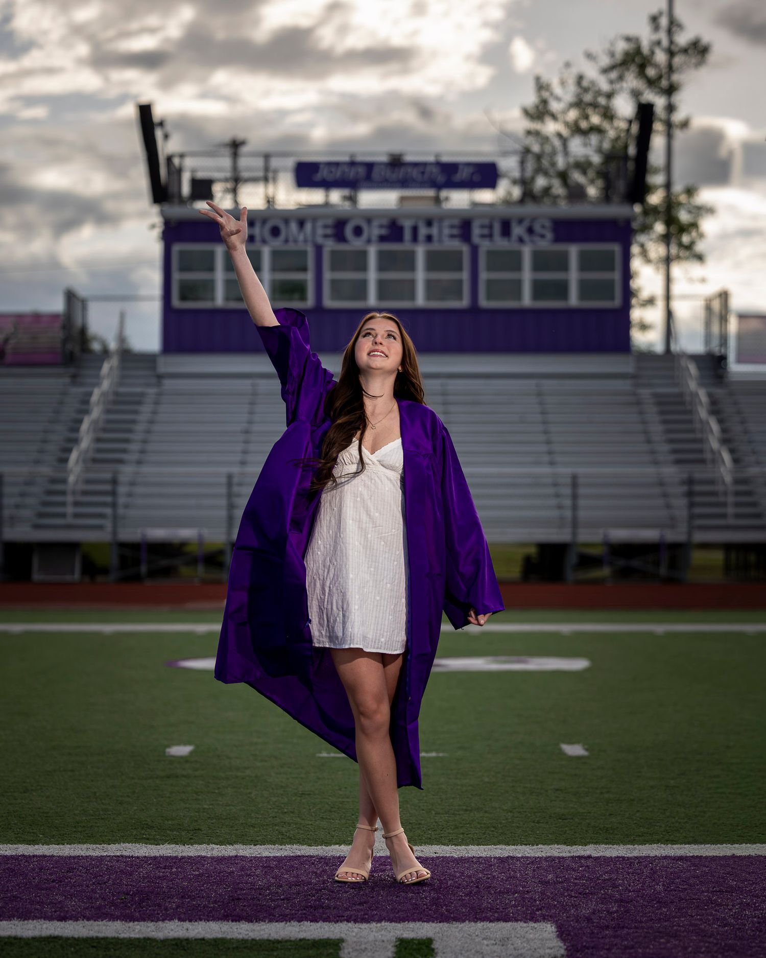Graduate in purple gown raises cap on football field at Home of the Elks stadium during senior portrait session.
