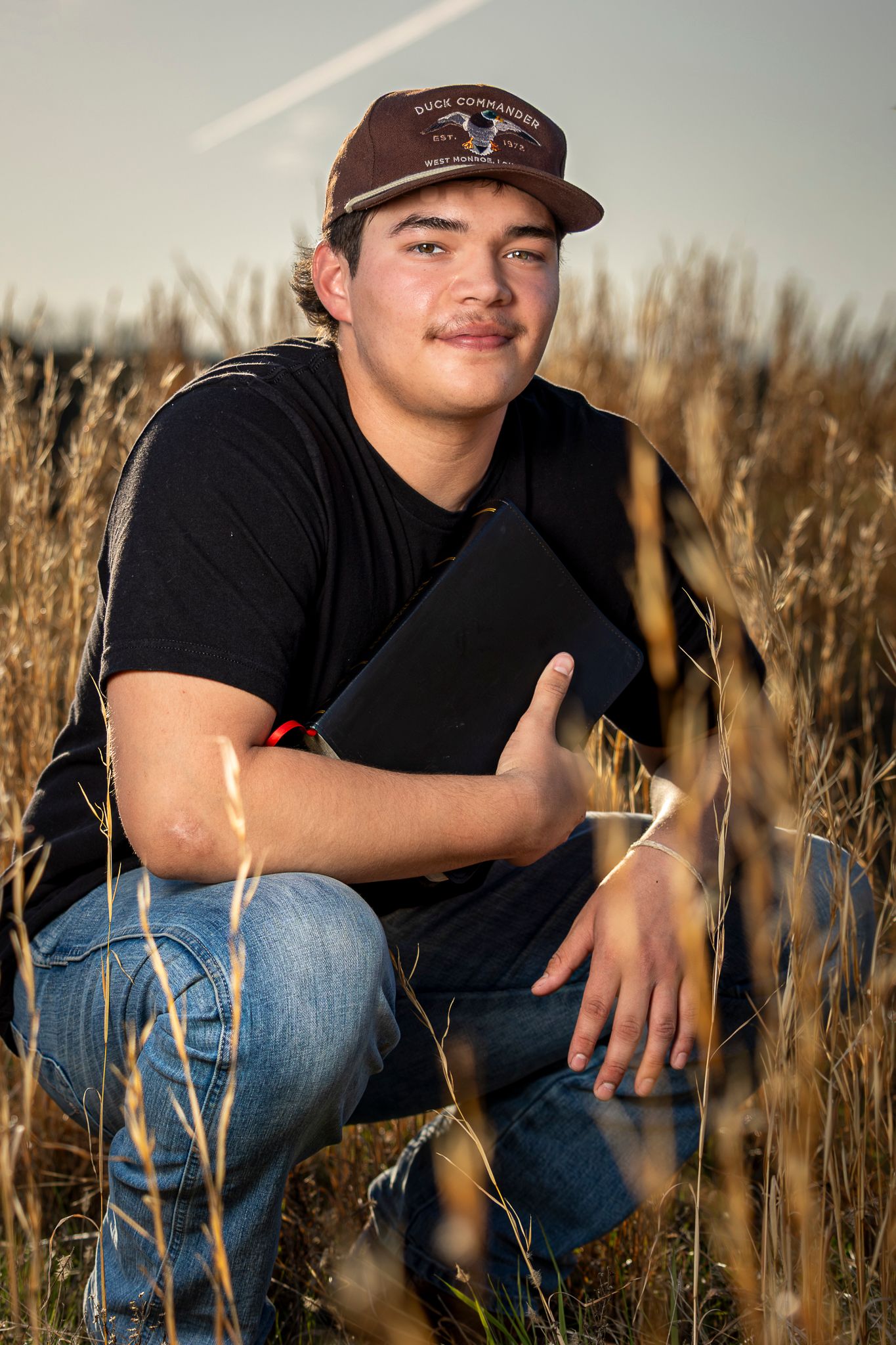 Teen boy in black shirt and cap crouching in wheat field holding a book.