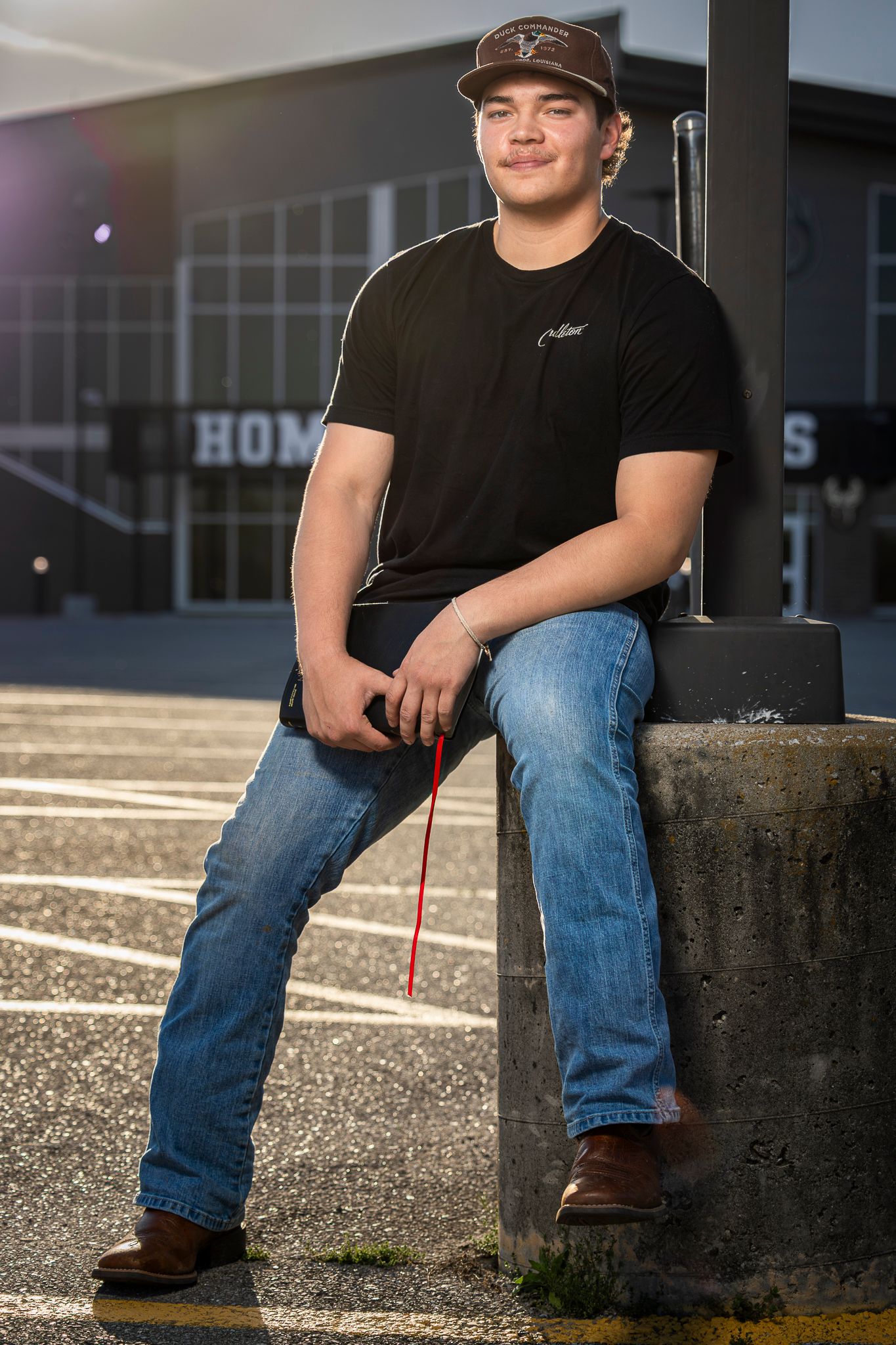 Young man in black t-shirt, jeans, and boots sits on concrete barrier in parking lot, smiling confidently.