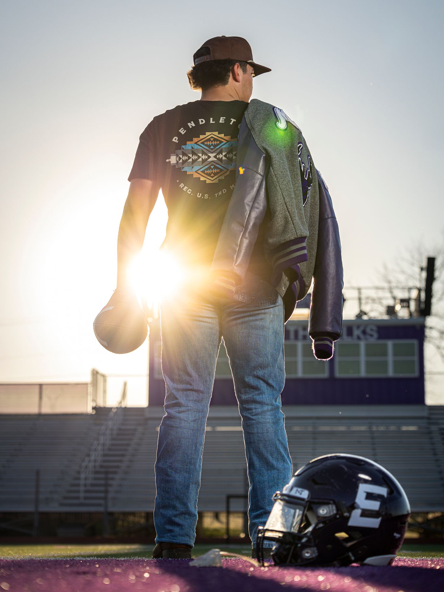 A young man stands on a football field at sunset, holding a helmet, wearing a Pendleton shirt and jeans.