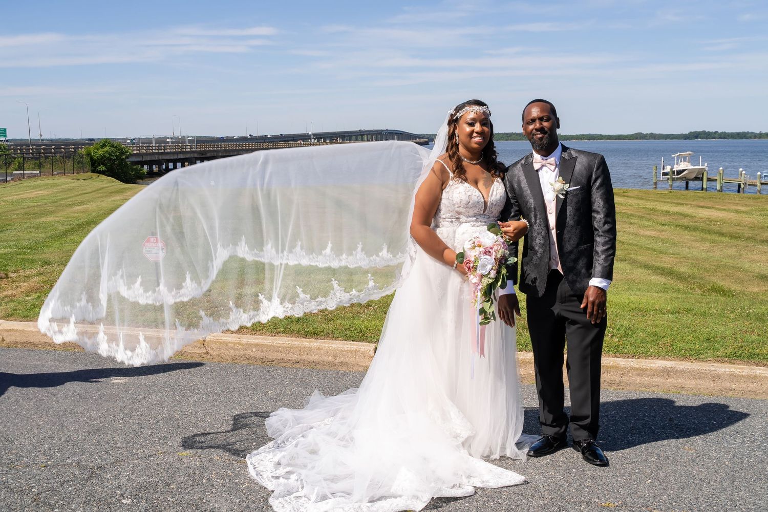 A happy couple poses on their wedding day with a flowing veil caught in the breeze by the waterfront.