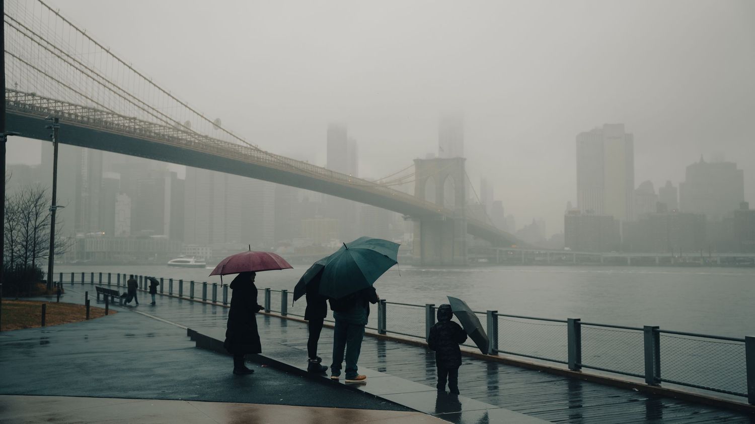 People with colorful umbrellas stand on a rainy waterfront promenade near the Brooklyn Bridge in foggy New York City.