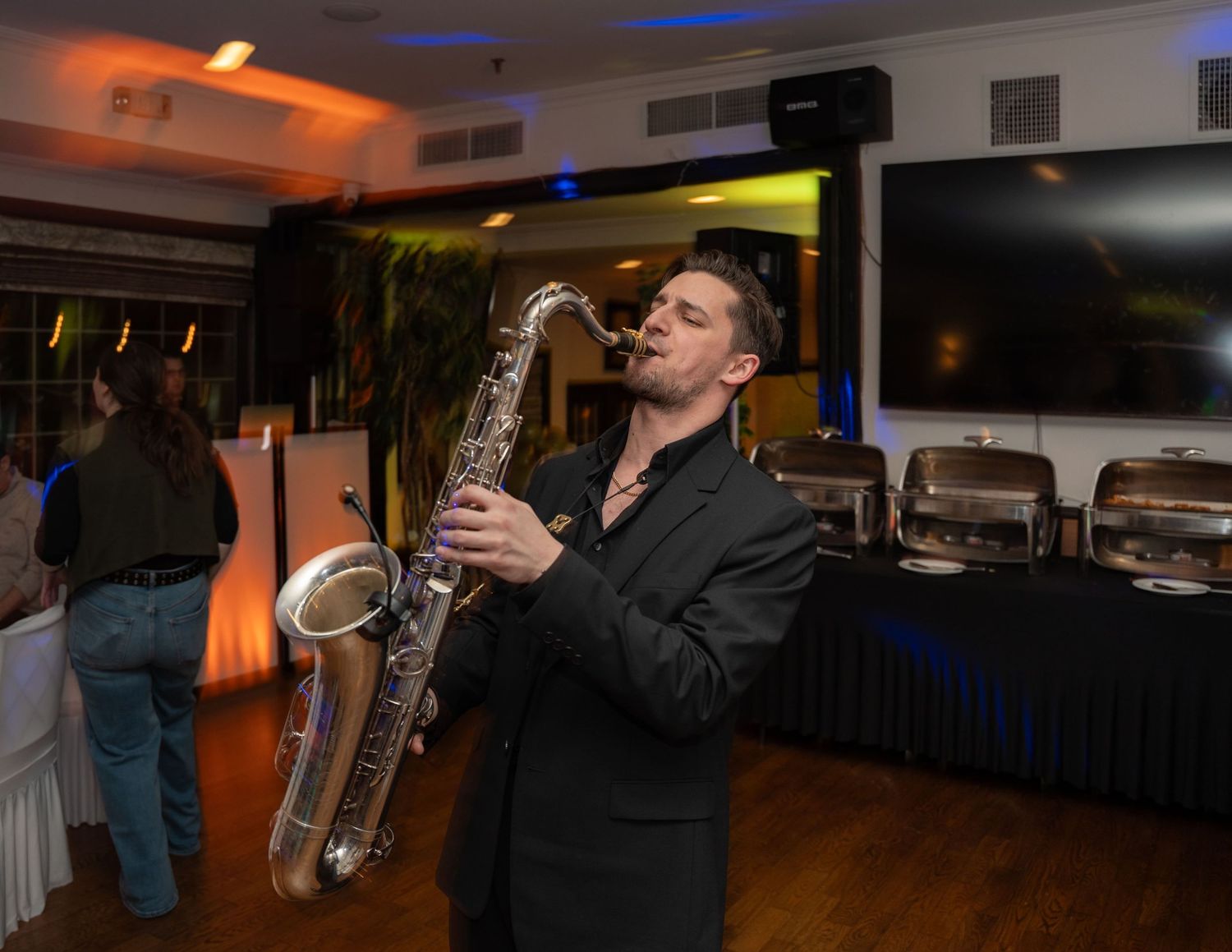 A man in a black suit plays saxophone at an elegant indoor event with chafing dishes and TV screens in the background.