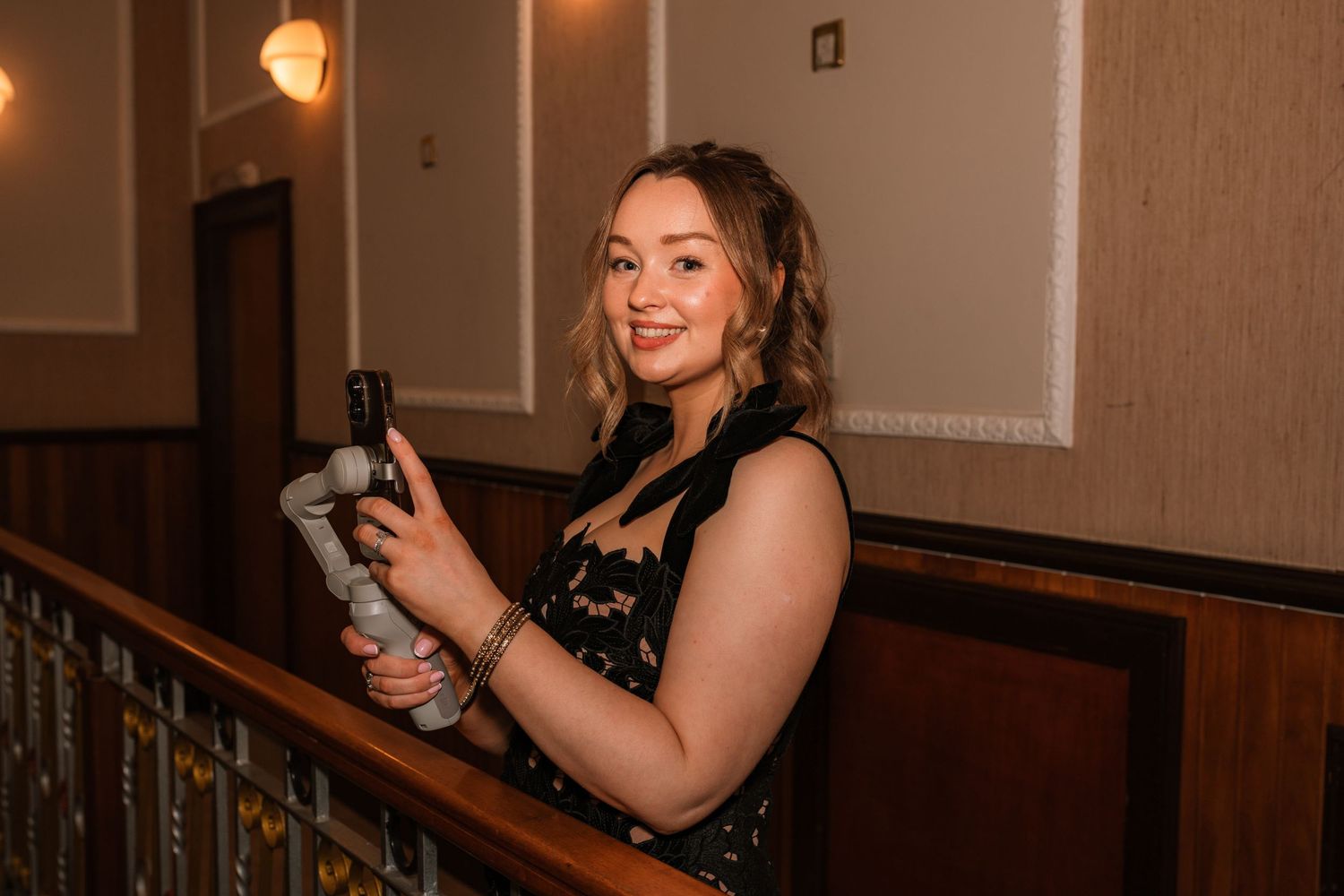 A person in a formal dress stands on a staircase while holding a mobile phone in a dimly lit setting.