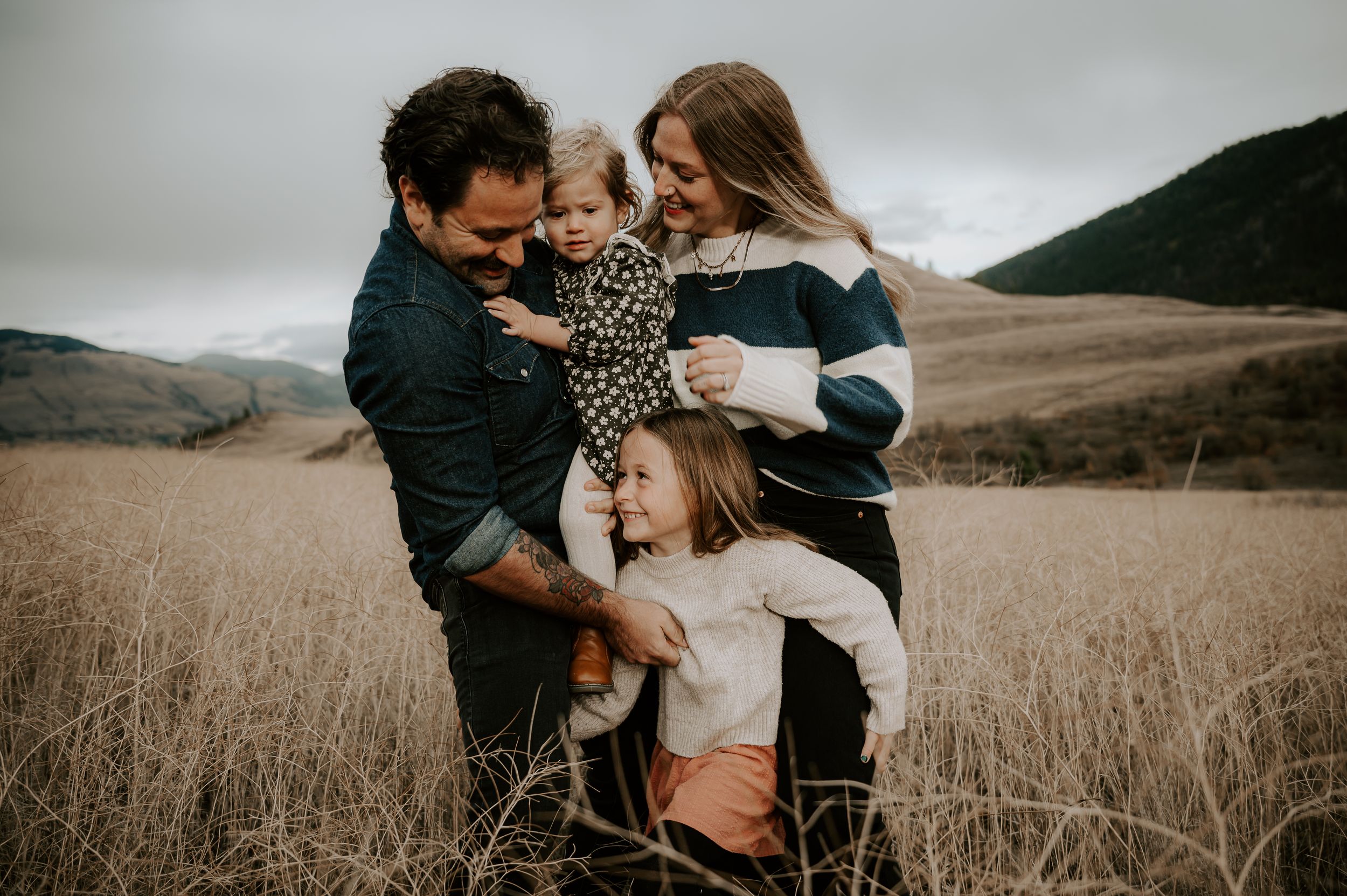 Happy family of four enjoying a scenic outdoor photo session in a golden field with misty mountains backdrop.