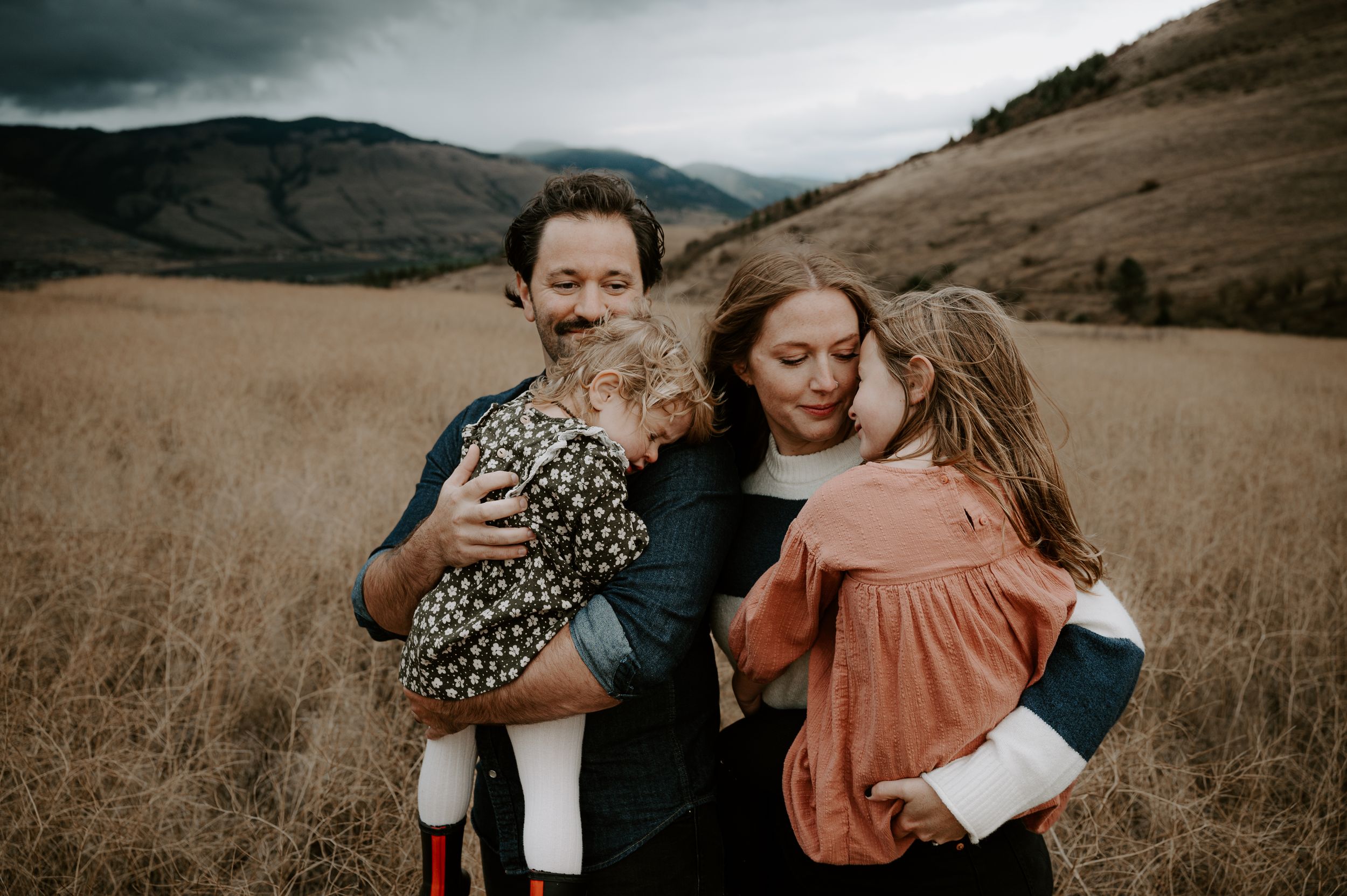 Family of four embracing in golden field with dramatic mountain backdrop during outdoor photo session.