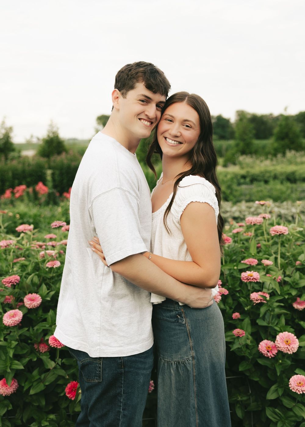 A smiling couple embraces among pink dahlia flowers in a lush garden, wearing casual white and dark outfits.