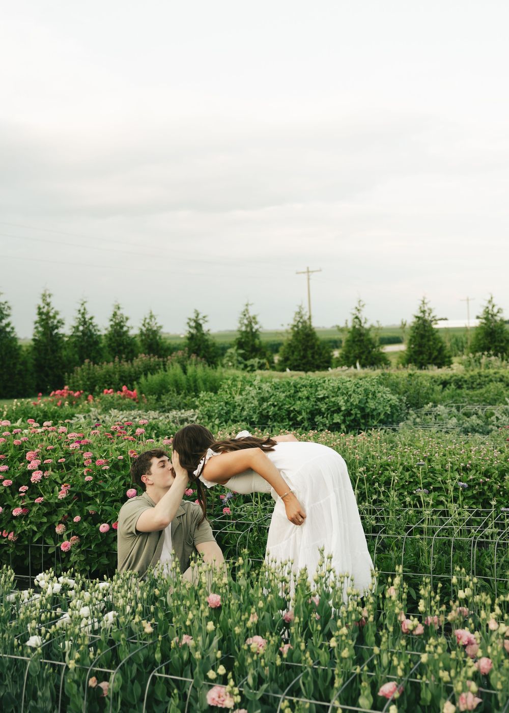 Couple sharing a romantic dip kiss in a lush flower garden with pink and white blooms under a cloudy sky.