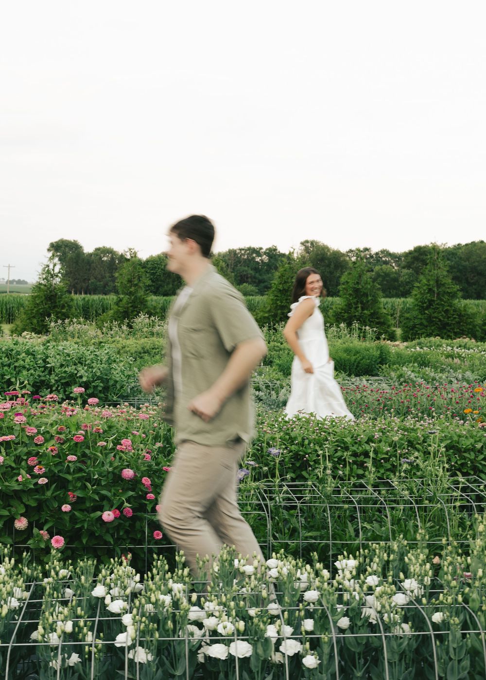 Man in olive shirt runs through colorful flower garden as woman in white dress smiles in background.