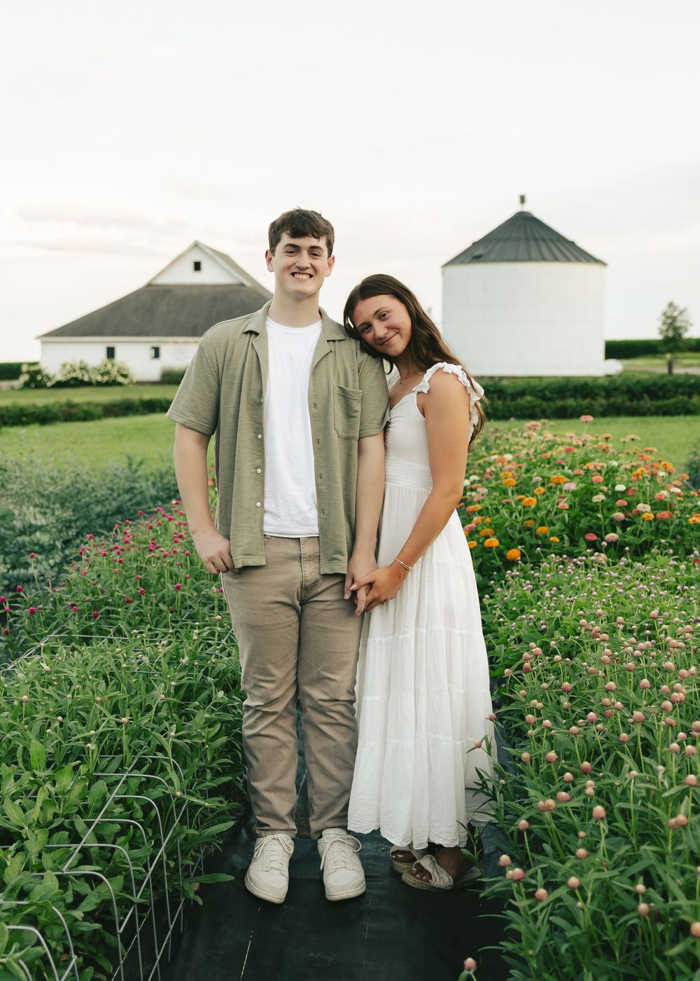 Couple posing in a colorful flower field with a white barn and silo in the background at sunset.