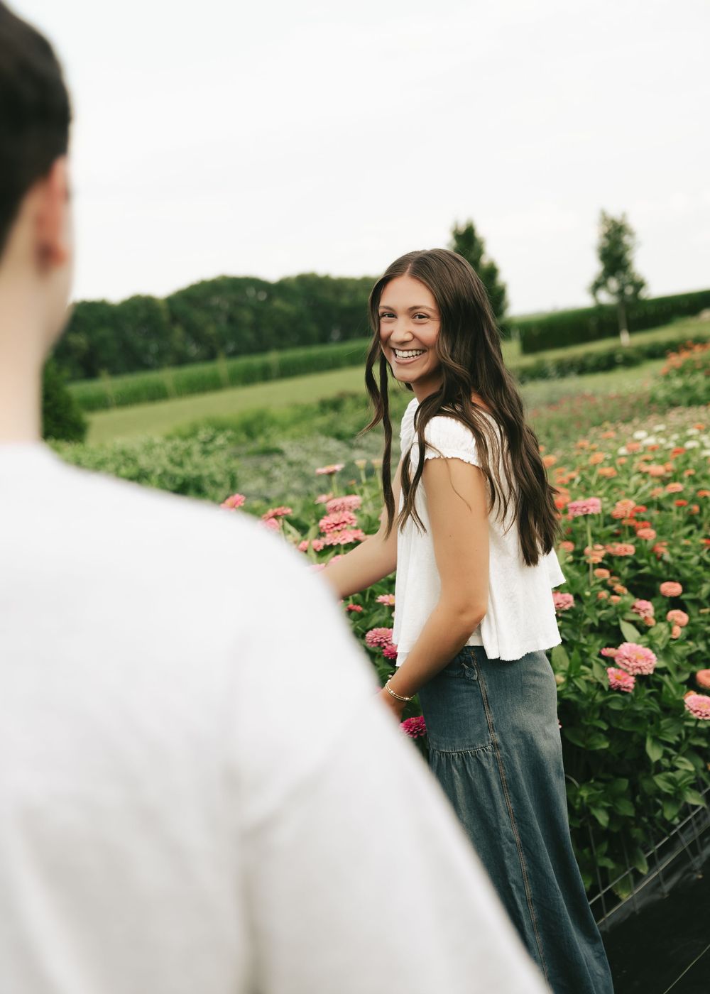 A smiling young woman with long dark hair stands among pink flowers in a garden, wearing a white top and jeans.