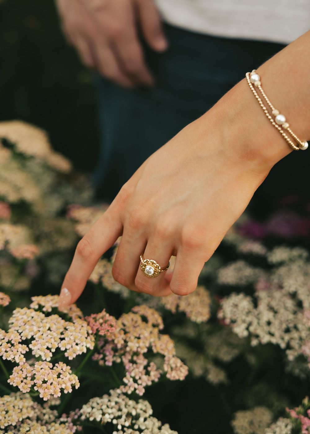 A woman's hand adorned with a gold gemstone ring and pearl bracelet delicately touches white wildflowers outdoors.
