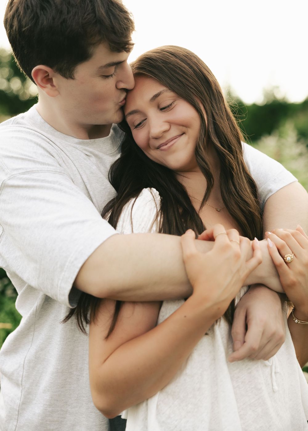 A young couple embraces outdoors, the man kissing her temple while she smiles, wearing an engagement ring.