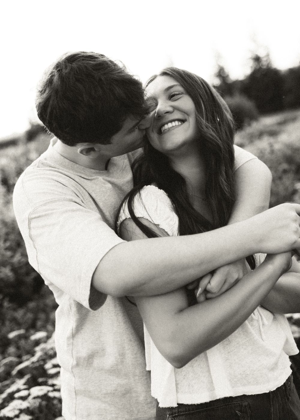 Black and white photo of a happy couple embracing outdoors, man kissing woman's cheek as she smiles joyfully.
