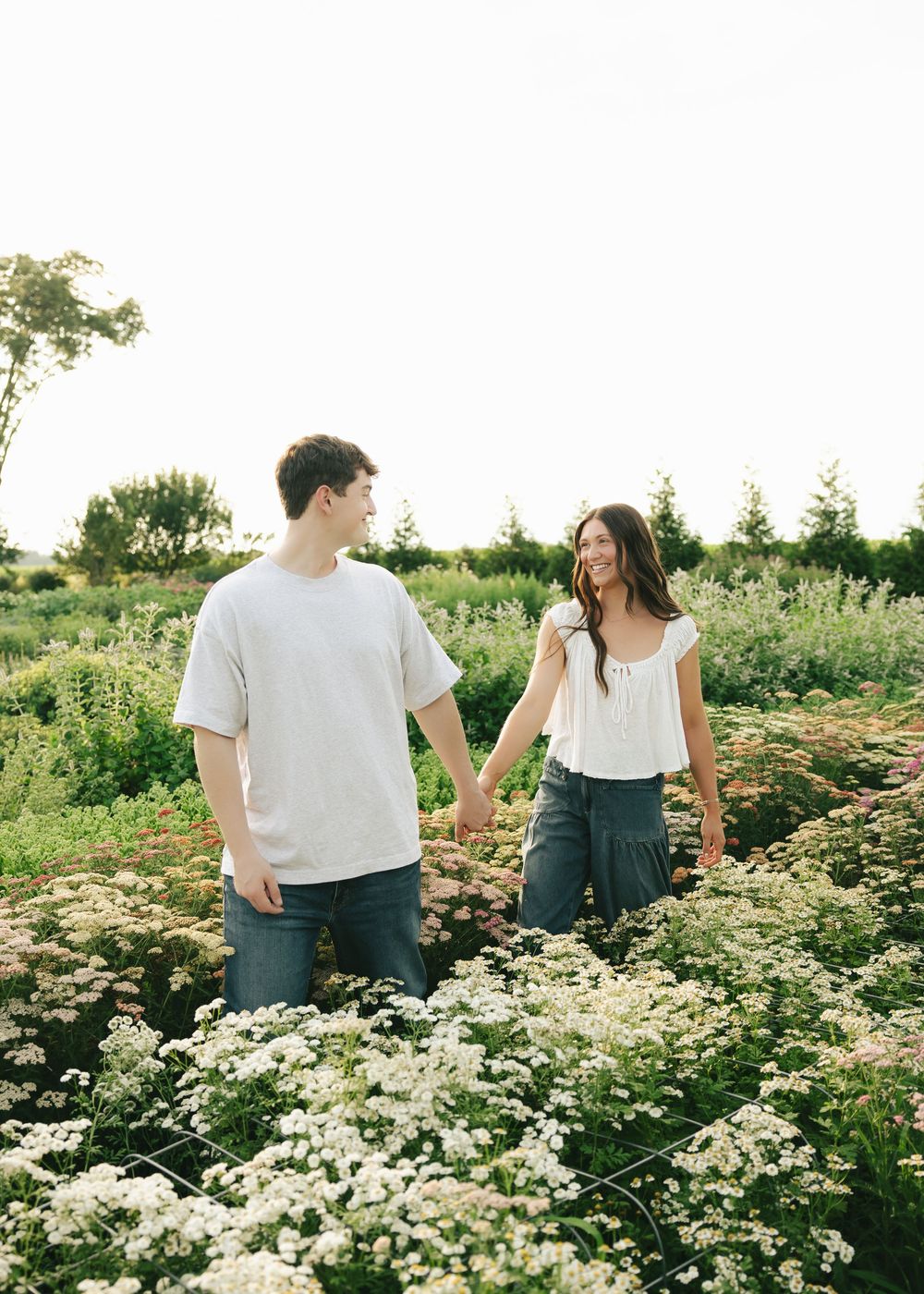 A couple holds hands while walking through a lush wildflower field, smiling at each other in golden hour light.