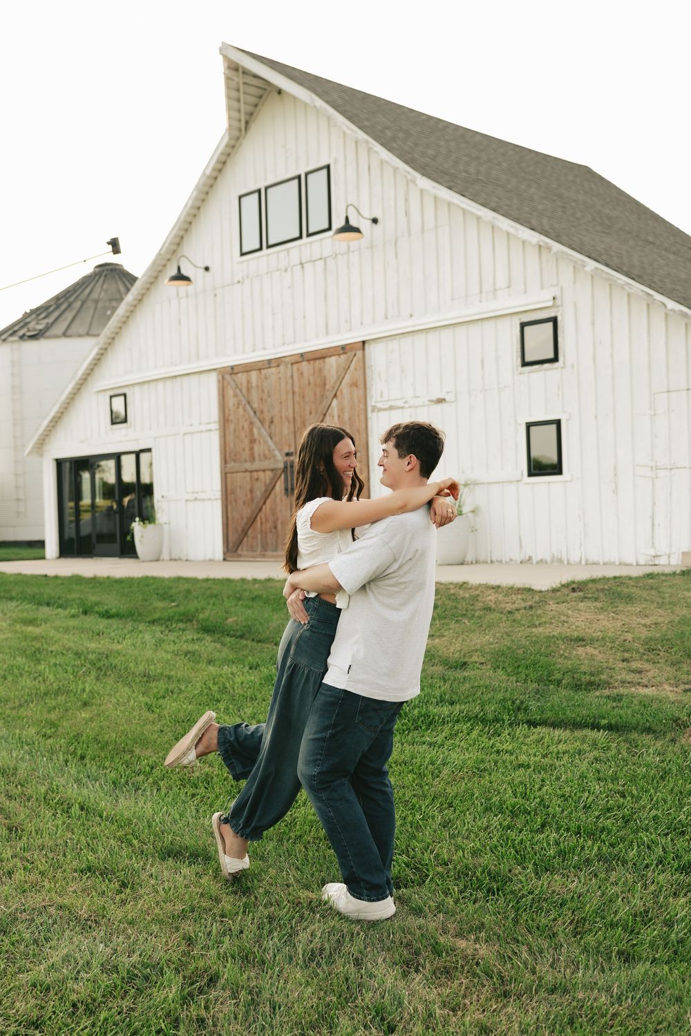 A joyful couple dancing and embracing on green grass in front of a charming white barn with wooden doors.