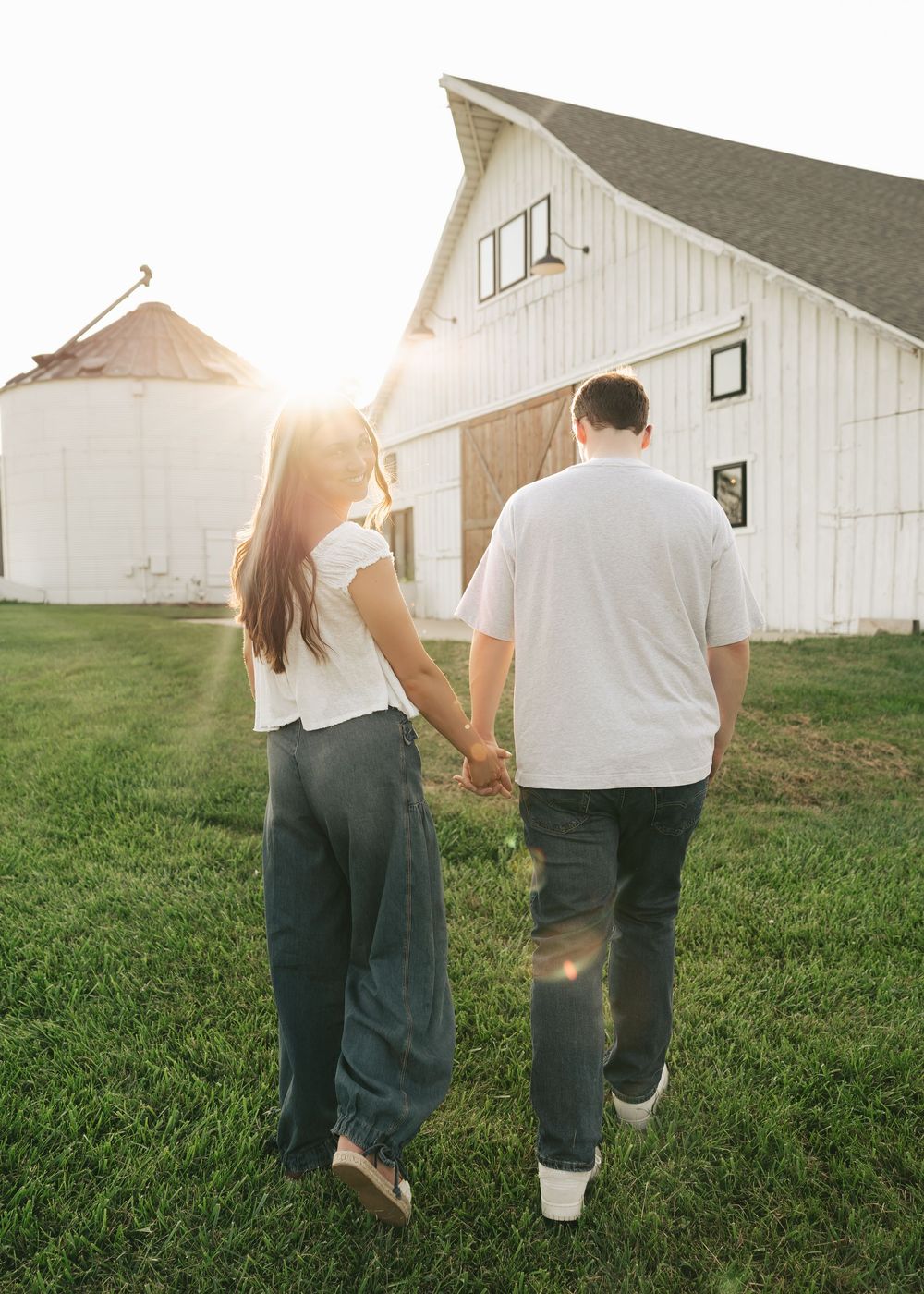 Couple holding hands walking toward a white barn at golden hour on a lush green farm.