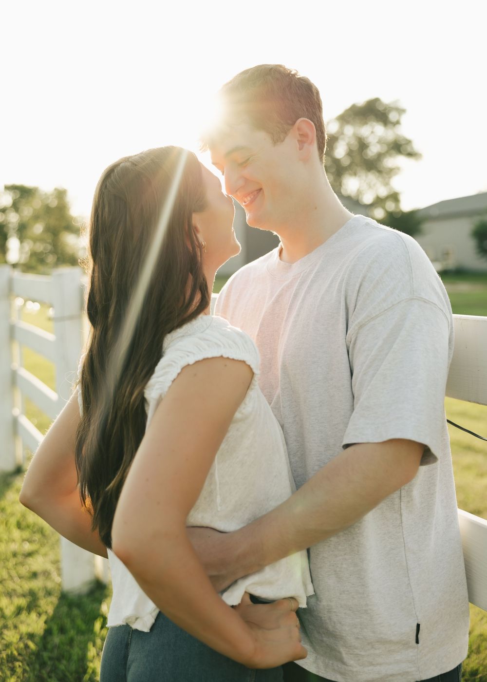 Couple sharing a romantic moment outdoors at golden hour near a white fence, sun glowing behind them.