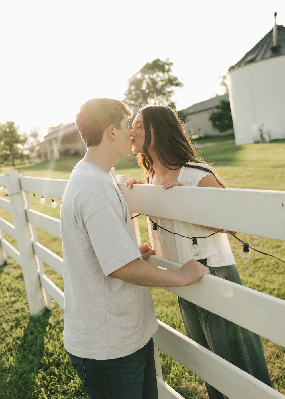Couple sharing a kiss by a white fence at a sunny farm during golden hour.