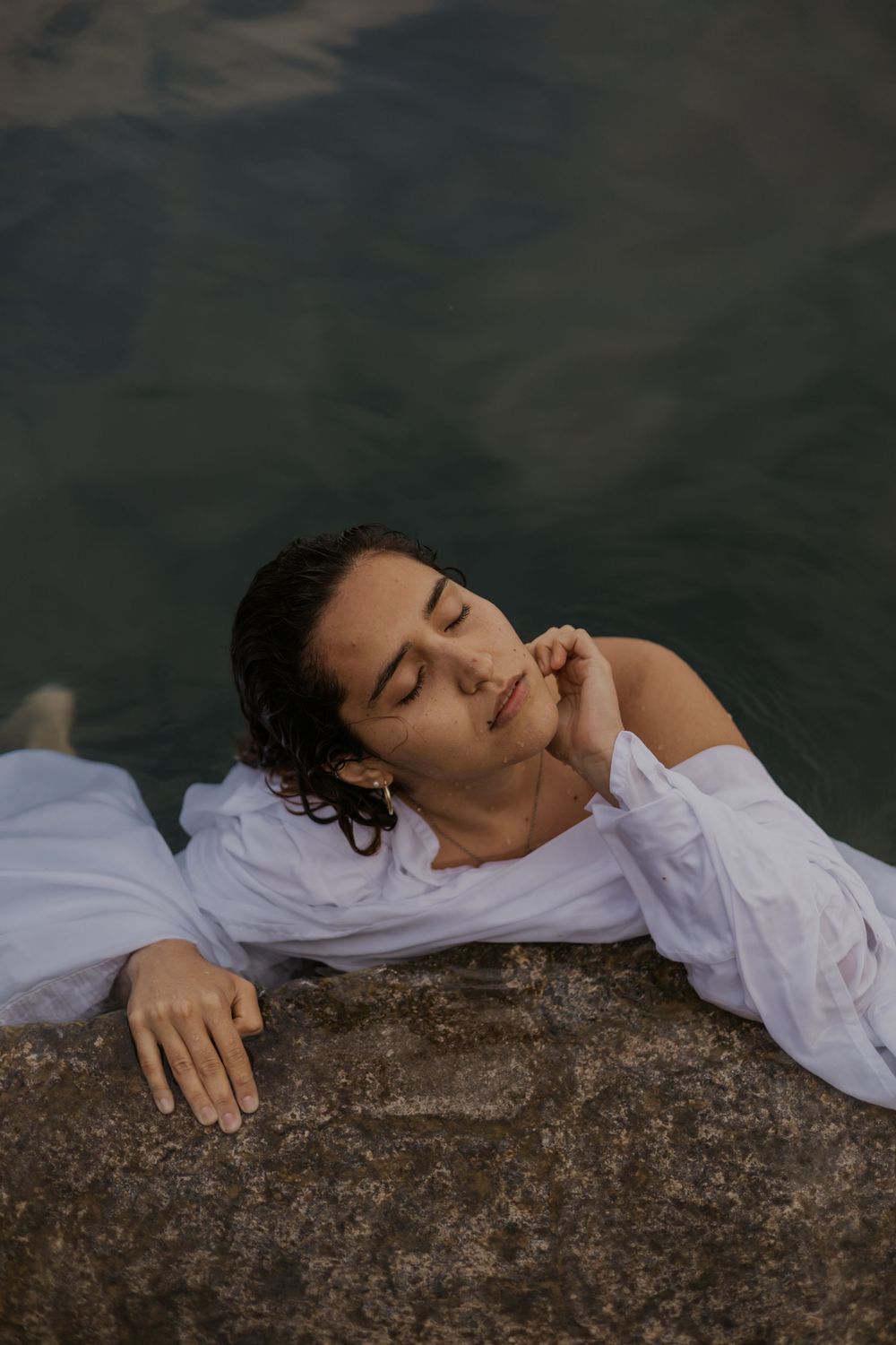Woman in white shirt resting peacefully on rocks by water with eyes closed.