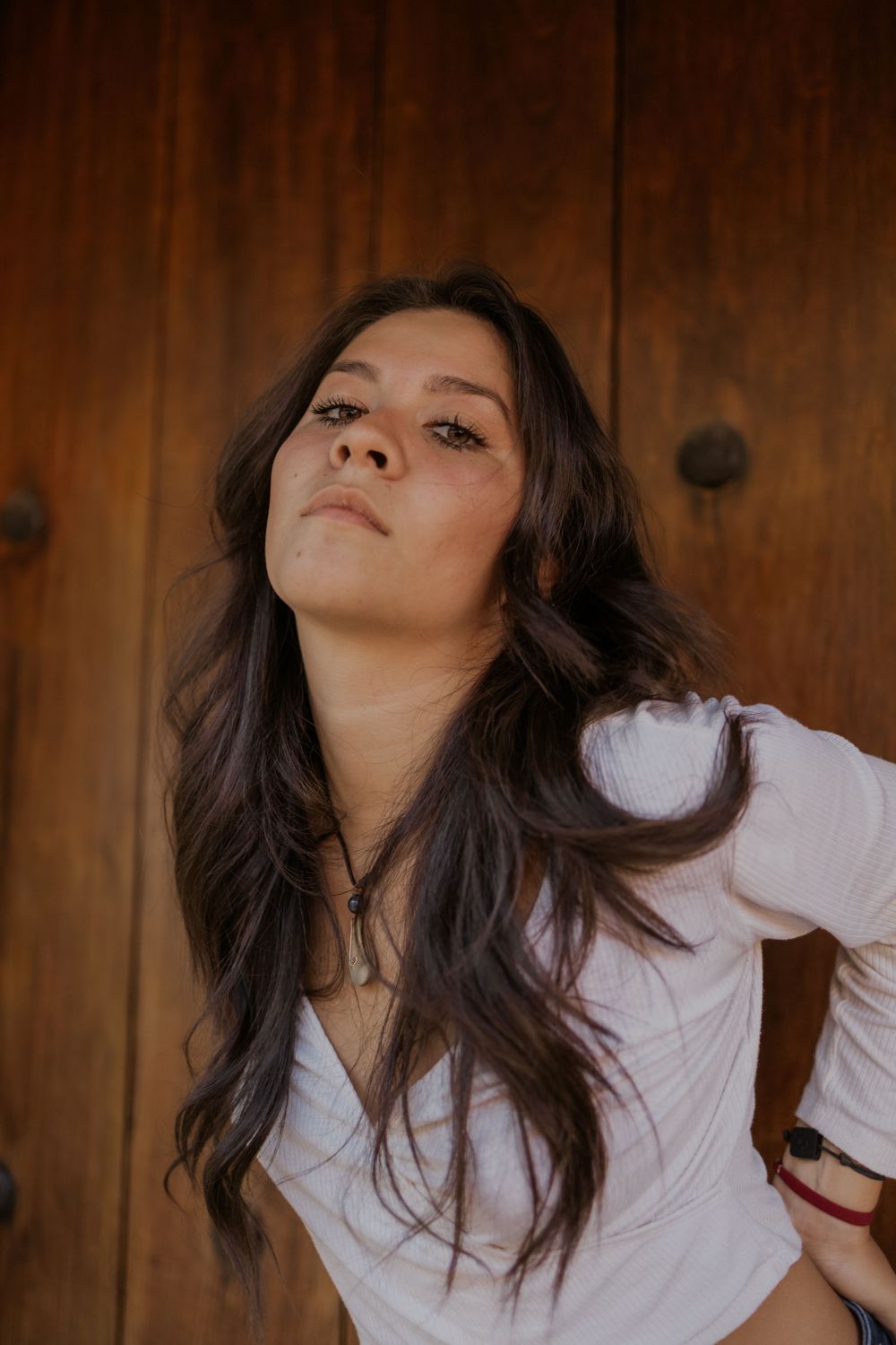 Young woman with long dark hair wearing a white top poses confidently against a rustic wooden door.