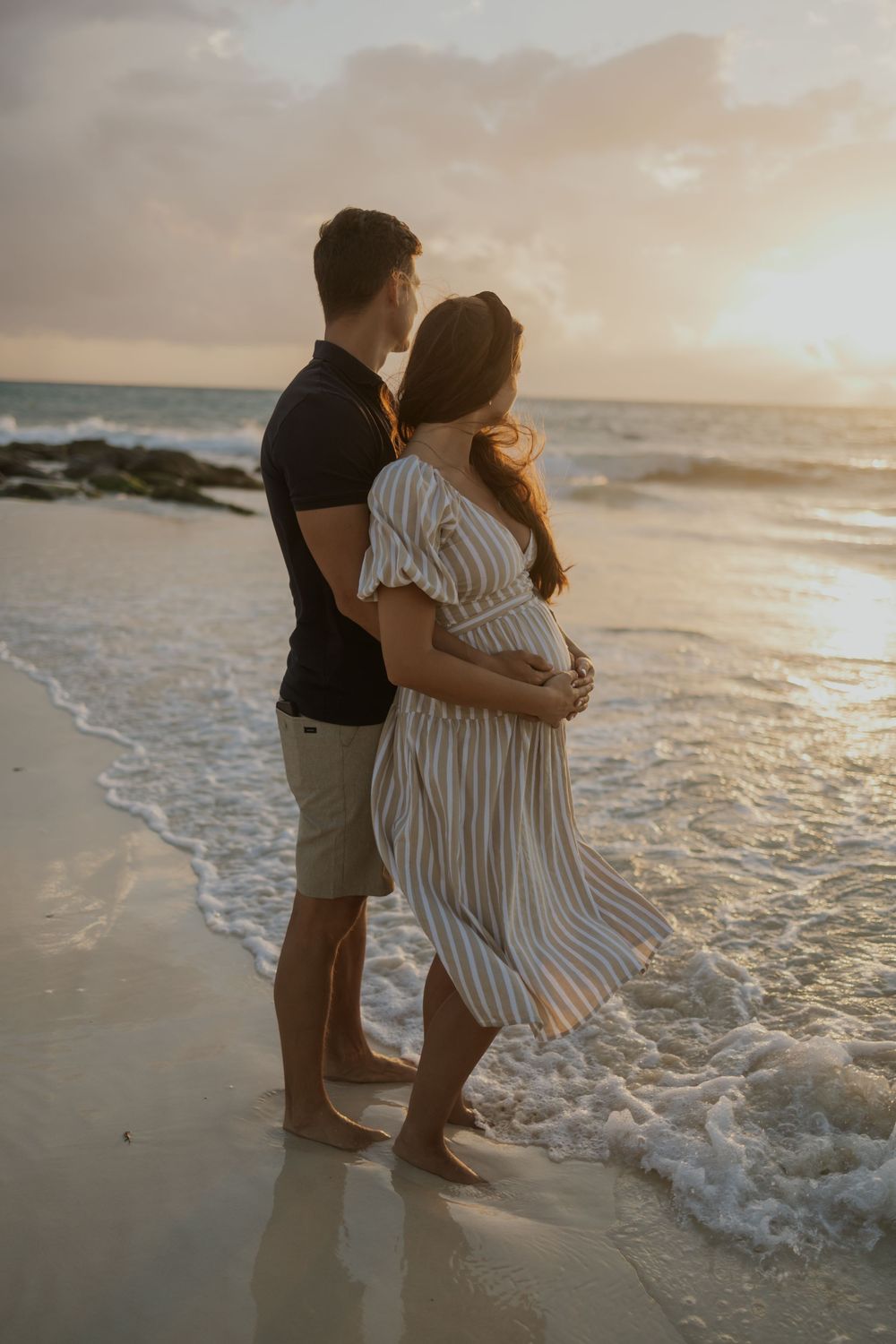 Pregnant couple embracing at sunset on a beach, waves washing over their feet.