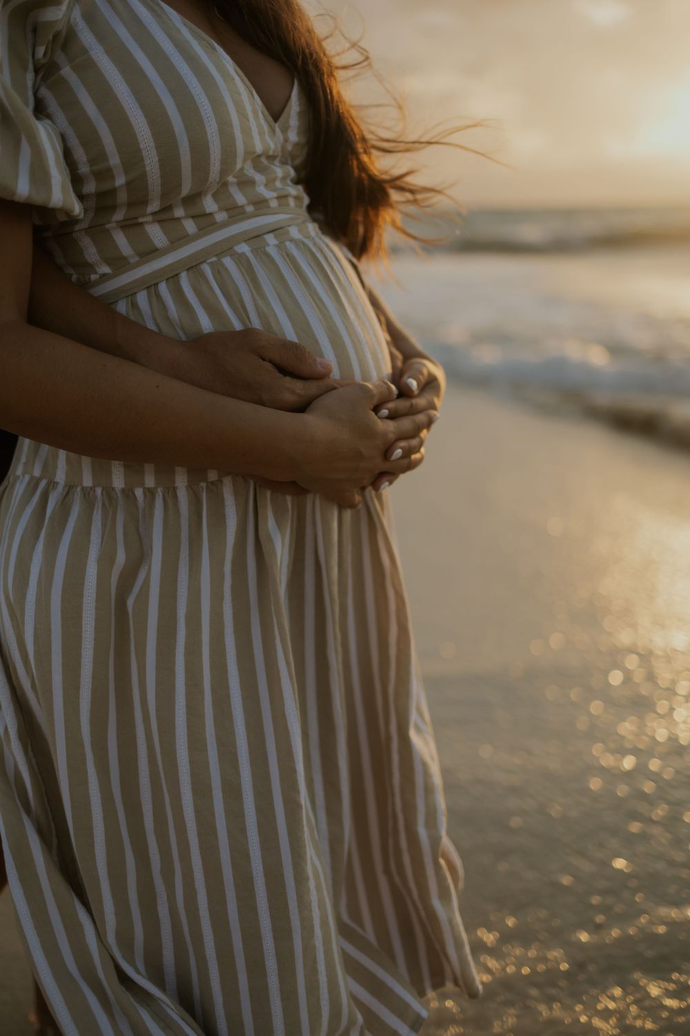Pregnant woman in striped dress cradling baby bump on beach at golden sunset.