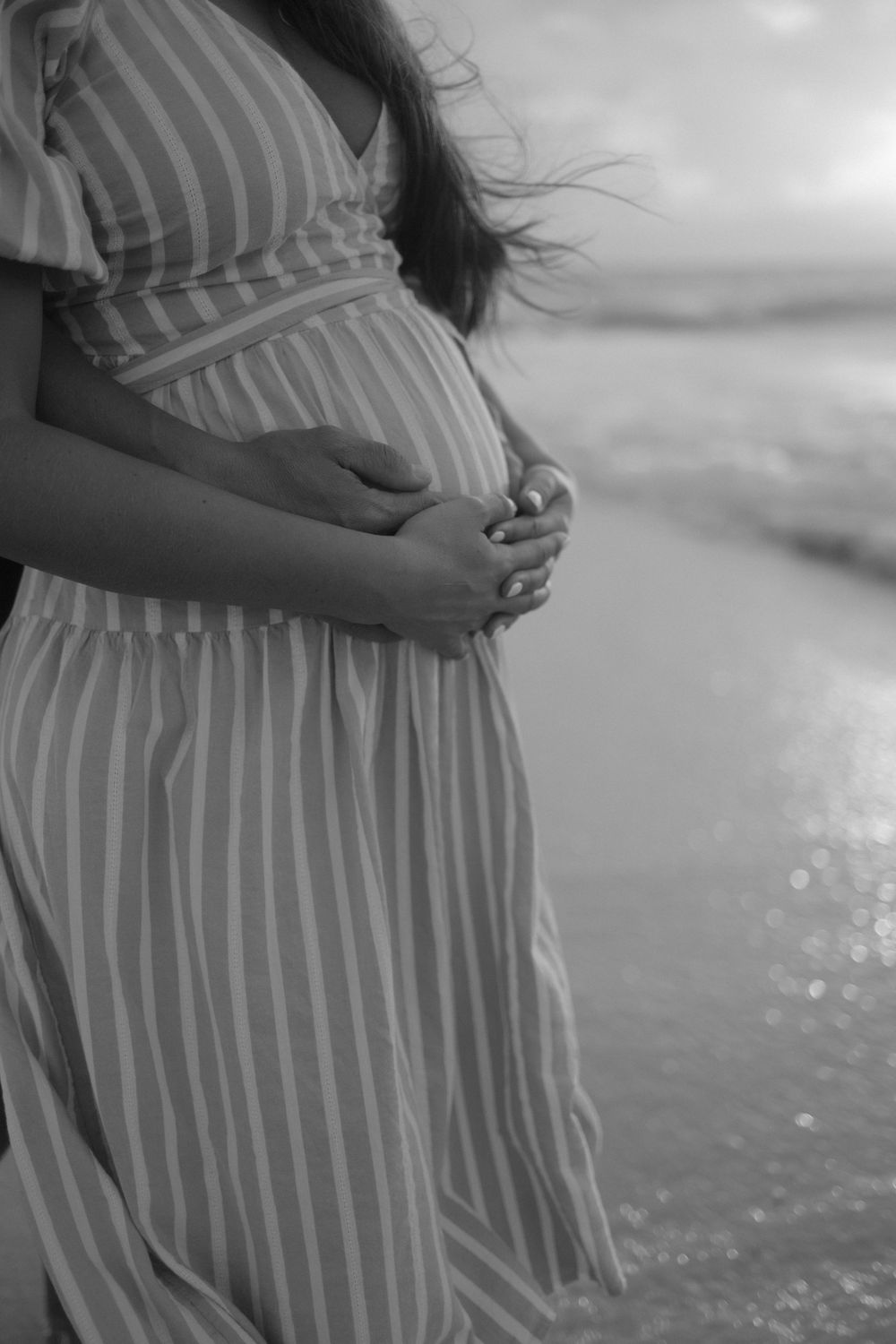 Black and white photo of a pregnant woman in a striped dress cradling her baby bump on a beach.