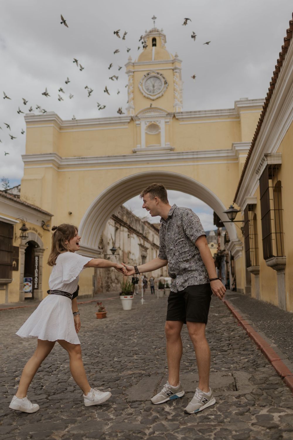 Couple holding hands and dancing in front of the Santa Catalina Arch in Antigua, Guatemala, with birds flying overhead.