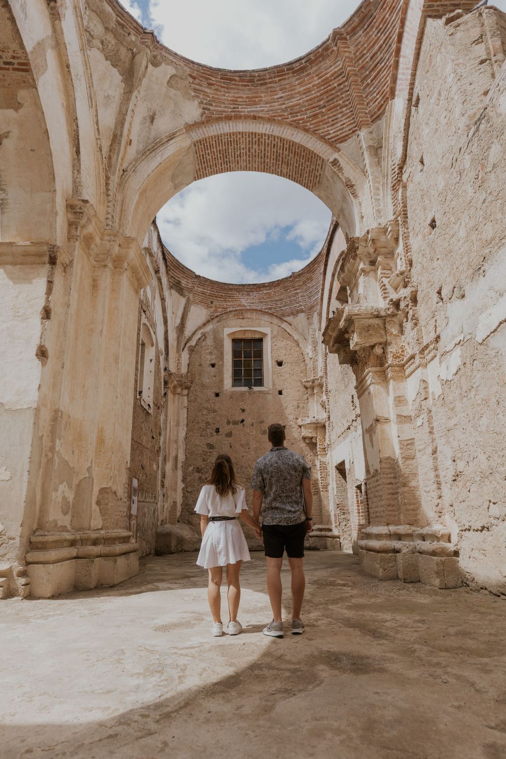 Couple holding hands exploring ancient ruins with grand arched ceilings and weathered stone walls.
