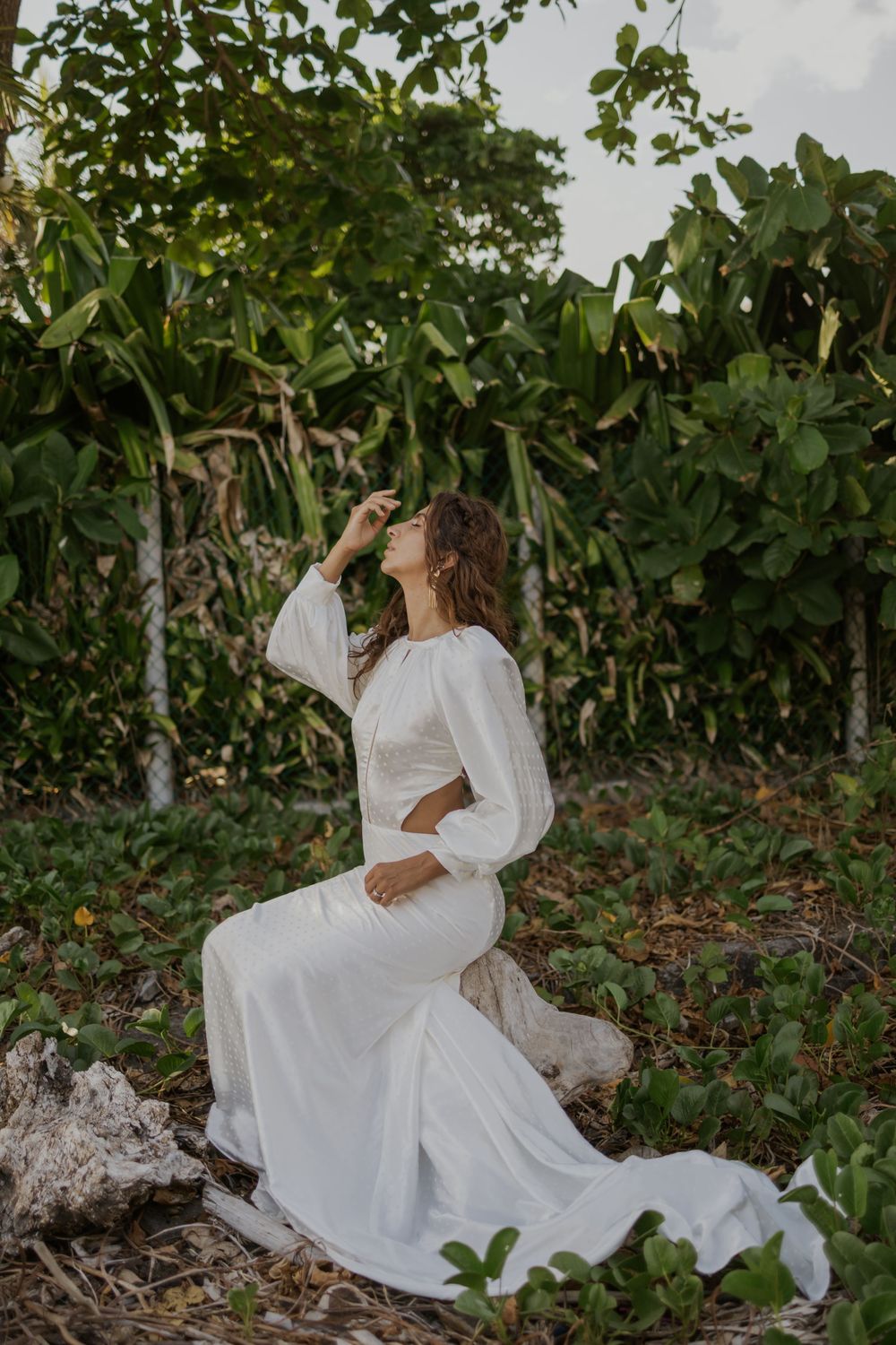 Woman in white boho wedding gown sitting gracefully among lush tropical greenery, gazing upward in serene outdoor setting.