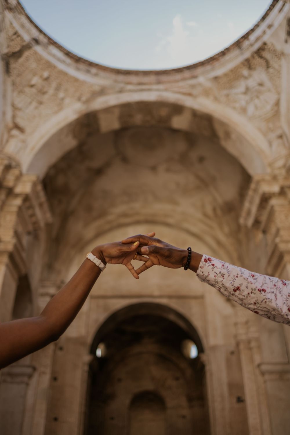 Two hands gently touching fingertips in front of a grand stone archway at an ancient historic landmark.