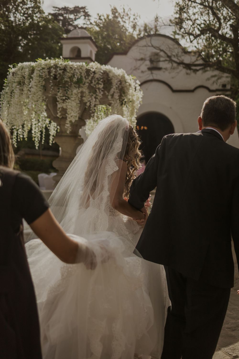 Bride in white gown and veil walking toward floral arch at outdoor wedding ceremony.