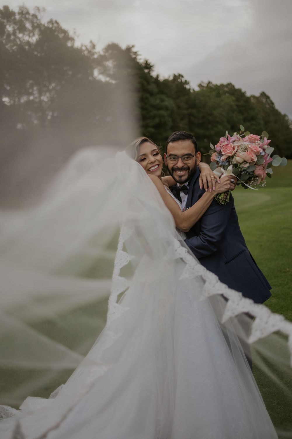 Joyful bride and groom embracing outdoors with flowing veil and pink bouquet on lush green golf course.