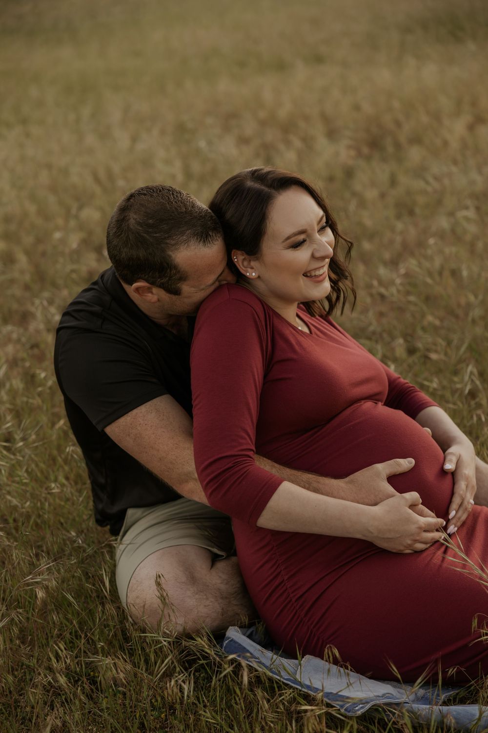 Pregnant woman in red dress sitting in a field with her partner kissing her cheek, both cradling her baby bump.