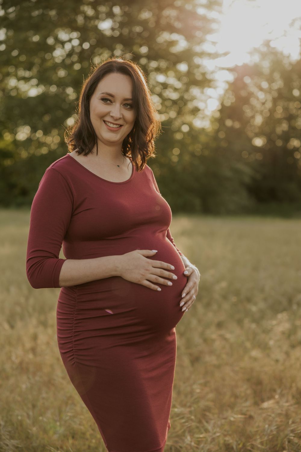 Smiling pregnant woman in burgundy dress cradling baby bump outdoors in golden sunlight.