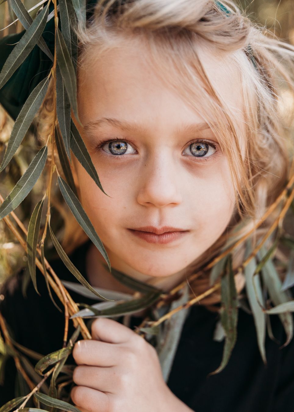 Portrait through eucalyptus leaves showing bright blue eyes and blonde hair in natural lighting.