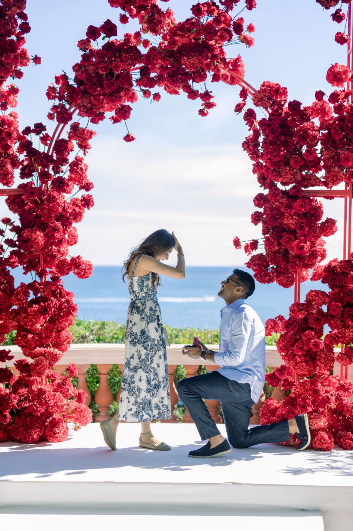 A couple sits beneath vibrant red bougainvillea flowers against a white wall with ocean views in the background.